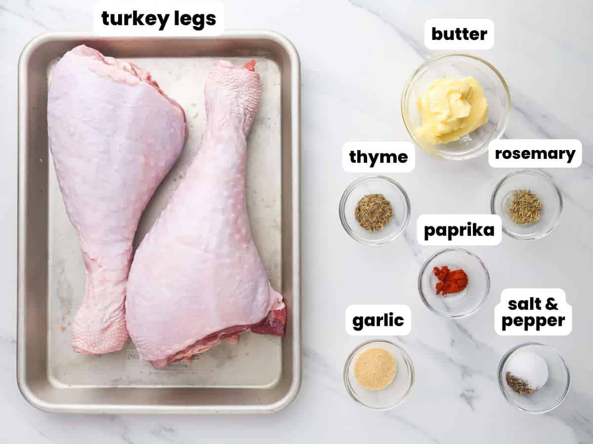 Two turkey legs on a small sheet pan. Next to the pan are small bowls filled with butter, herbs, and seasonings. Each ingredient is labeled