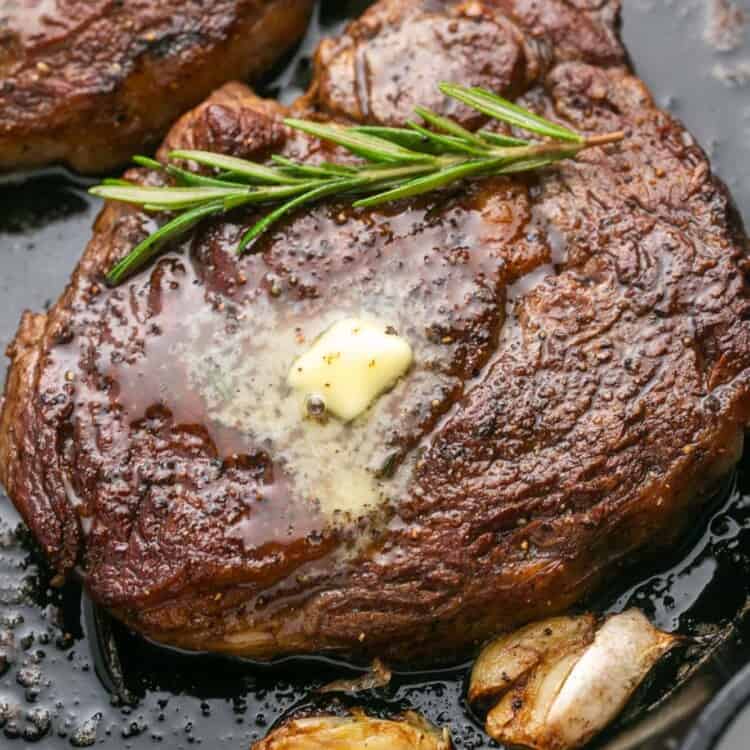 ribeye steak in a cast iron pan with herbs, butter, and garlic cloves