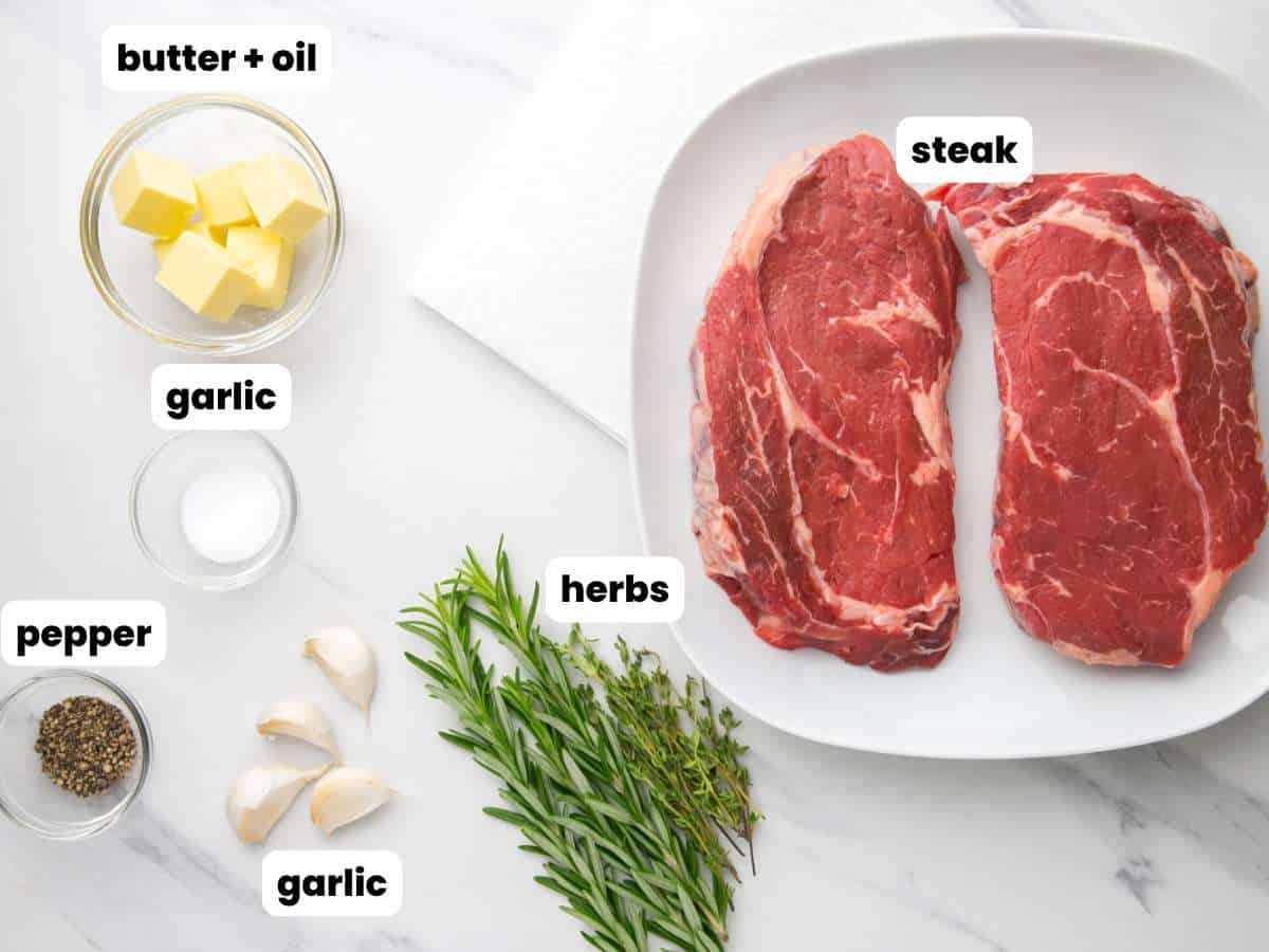 Two ribeye steaks on a white plate. Next to the plate are fresh herbs, butter, garlic, salt, and pepper