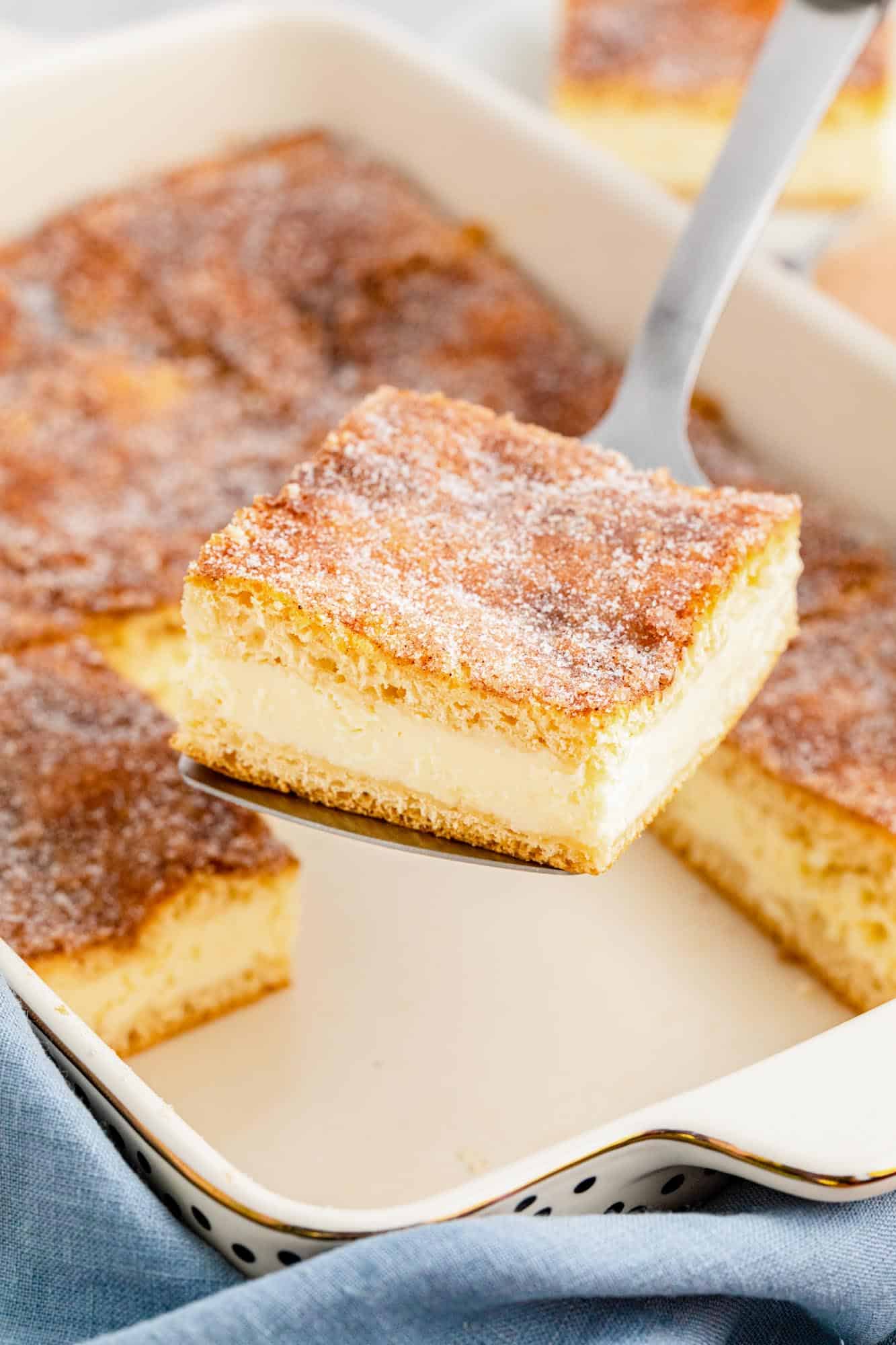 a ceramic 9 x 13 dish with sopapilla cheesecake bars. A spatula is lifting one out of the pan. Two other pieces have already been removed.