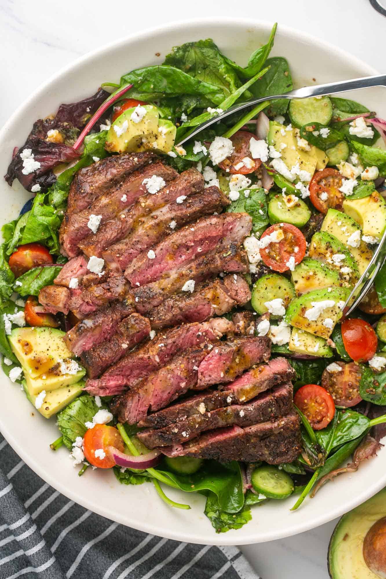 a salad with avocado and crumbled cheese, topped with sliced steak and dressing, in a large serving bowl.
