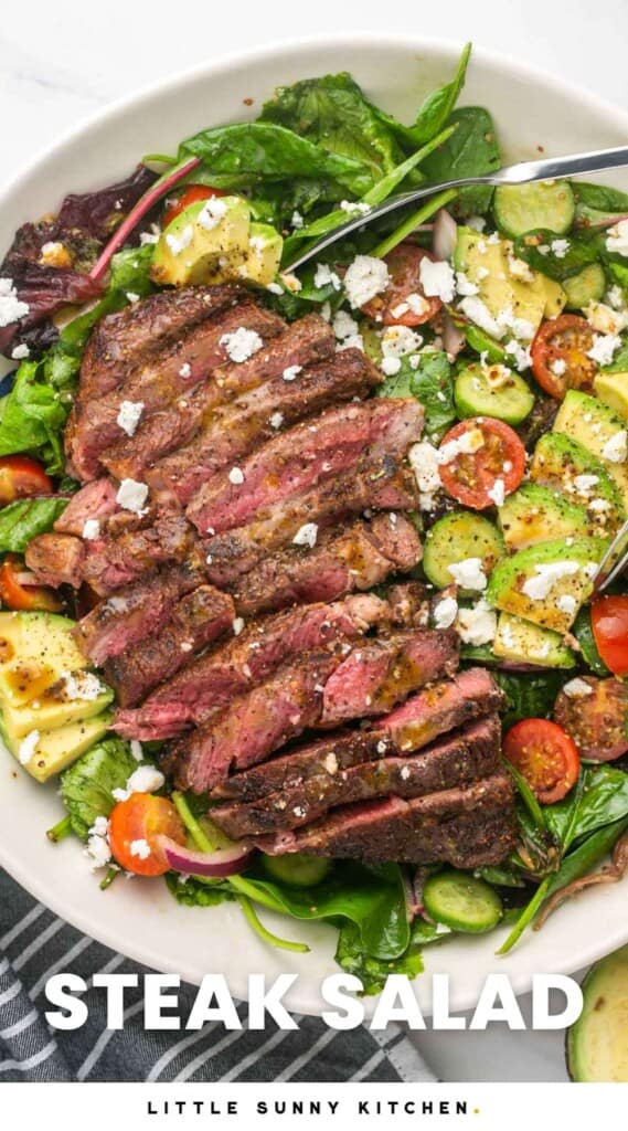 a salad with avocado and crumbled cheese, topped with sliced steak and dressing, in a large serving bowl.