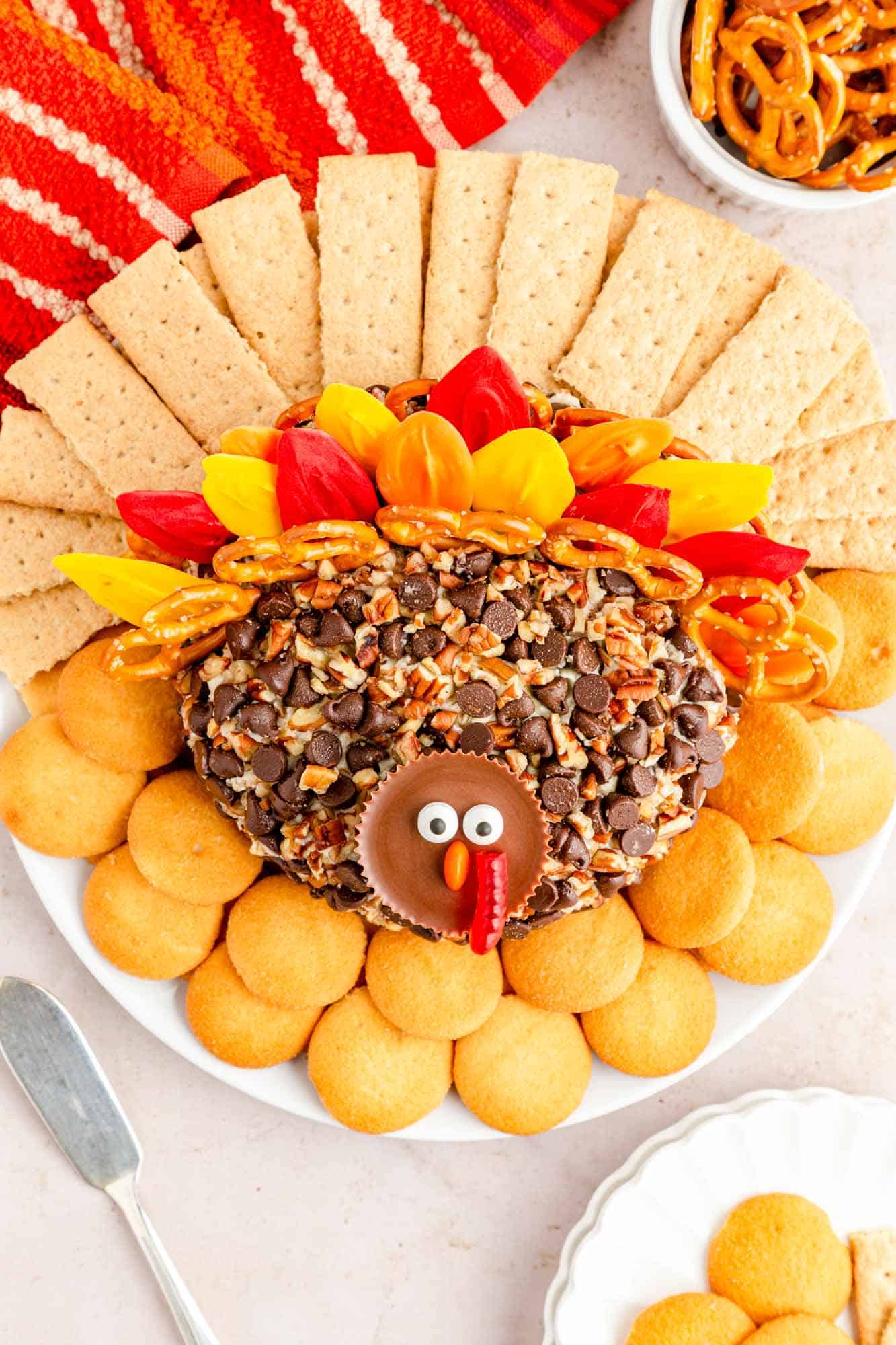 dessert turkey cheese ball viewed from above. It has a face made with a peanut butter cup, colorful candy feathers, and is surrounded by nilla wafers and graham crackers.