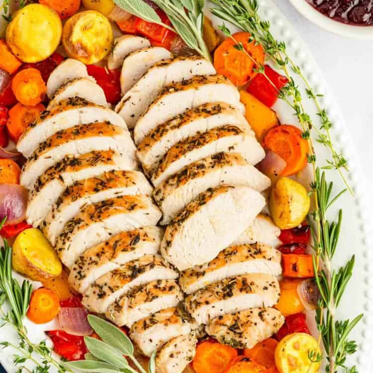 Overhead shot of sliced turkey tenderloin in a large oval white platter, with roasted vegetables and fresh herbs.