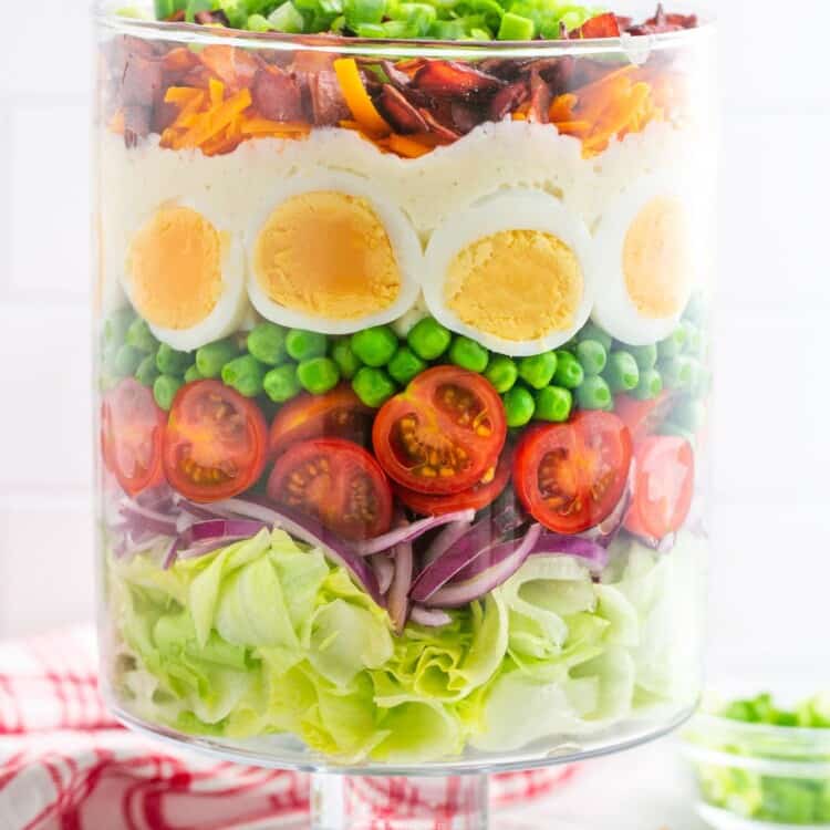 A tall clear glass trifle dish with a layered salad in it. Iceberg lettuce is the bottom layer, then sliced red onion, cherry tomatoes, peas, hard boiled eggs, cheeses, bacon, and green onion