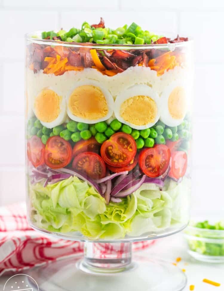 A tall clear glass trifle dish with a layered salad in it. Iceberg lettuce is the bottom layer, then sliced red onion, cherry tomatoes, peas, hard boiled eggs, cheeses, bacon, and green onion