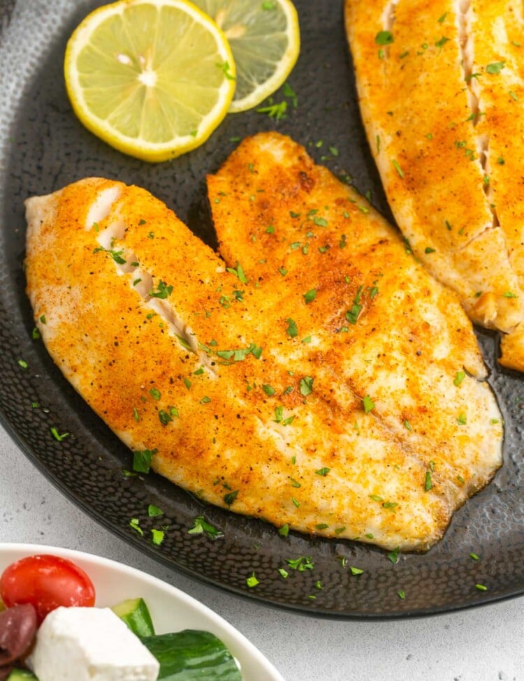 Overhead shot of air fried tilapia fillet on a black plate, and lemon slices on the side