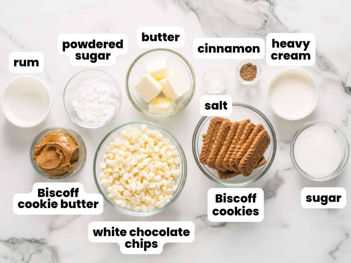 The ingredients you need to make a chocolate salami with white chocolate and biscoff cookies, all in separate bowls on a marble counter. Text boxes label each bowl.