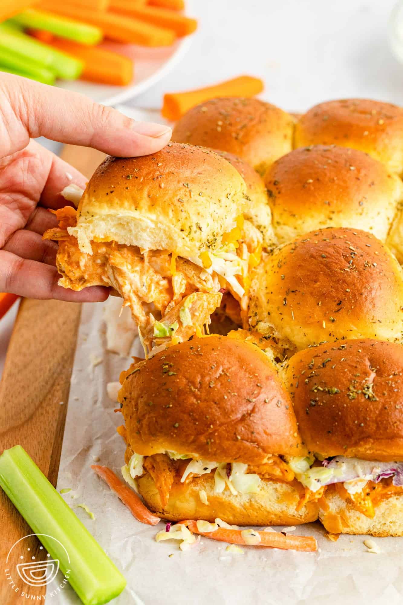 a hand pulling a buffalo chicken slider away from the rest. The tops of the sandwiches are golden brown. Celery and carrot sticks are on a plate in the background.