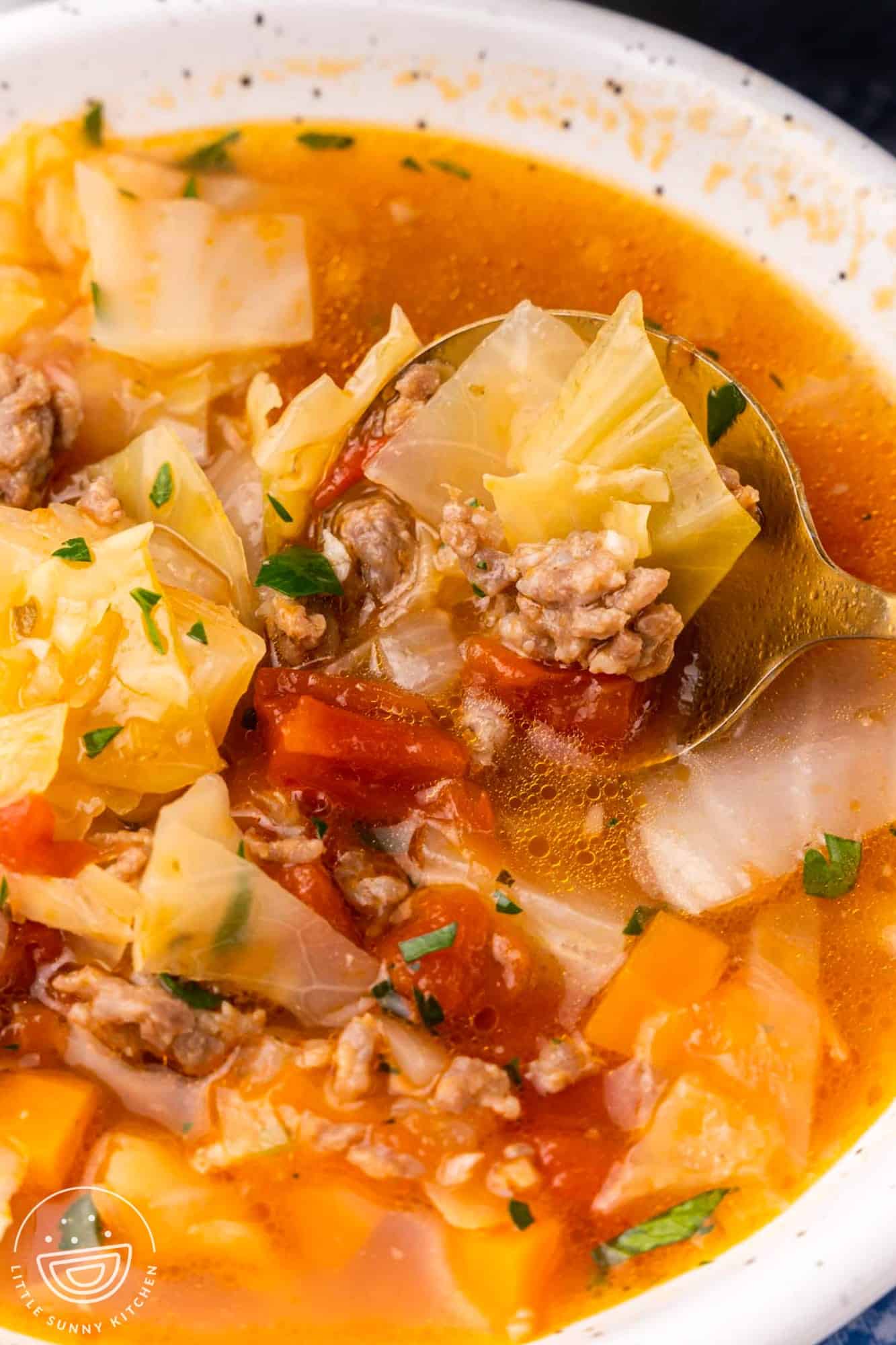 A white bowl of cabbage and sausage soup with tomatoes. A spoon is lifting up a bite.