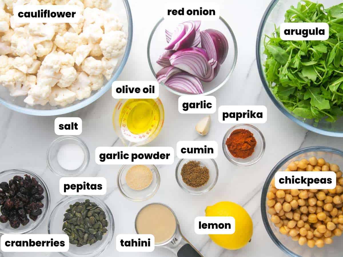 Ingredients needed to make a roasted cauliflower salad including cauliflower florets, onion, arugula, tahini, chickpeas, and seasonings.