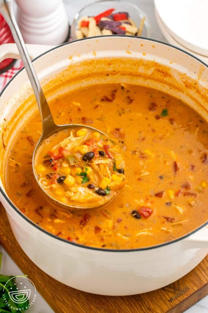 a white dutch oven of creamy chicken enchilada soup. a ladle is lifting soup out of the pot