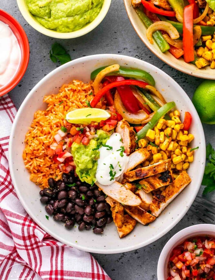 a white bowl filled with mexican rice, sliced chicken, fajita veggies, corn, and beans. Topped with sour cream and guacamole.