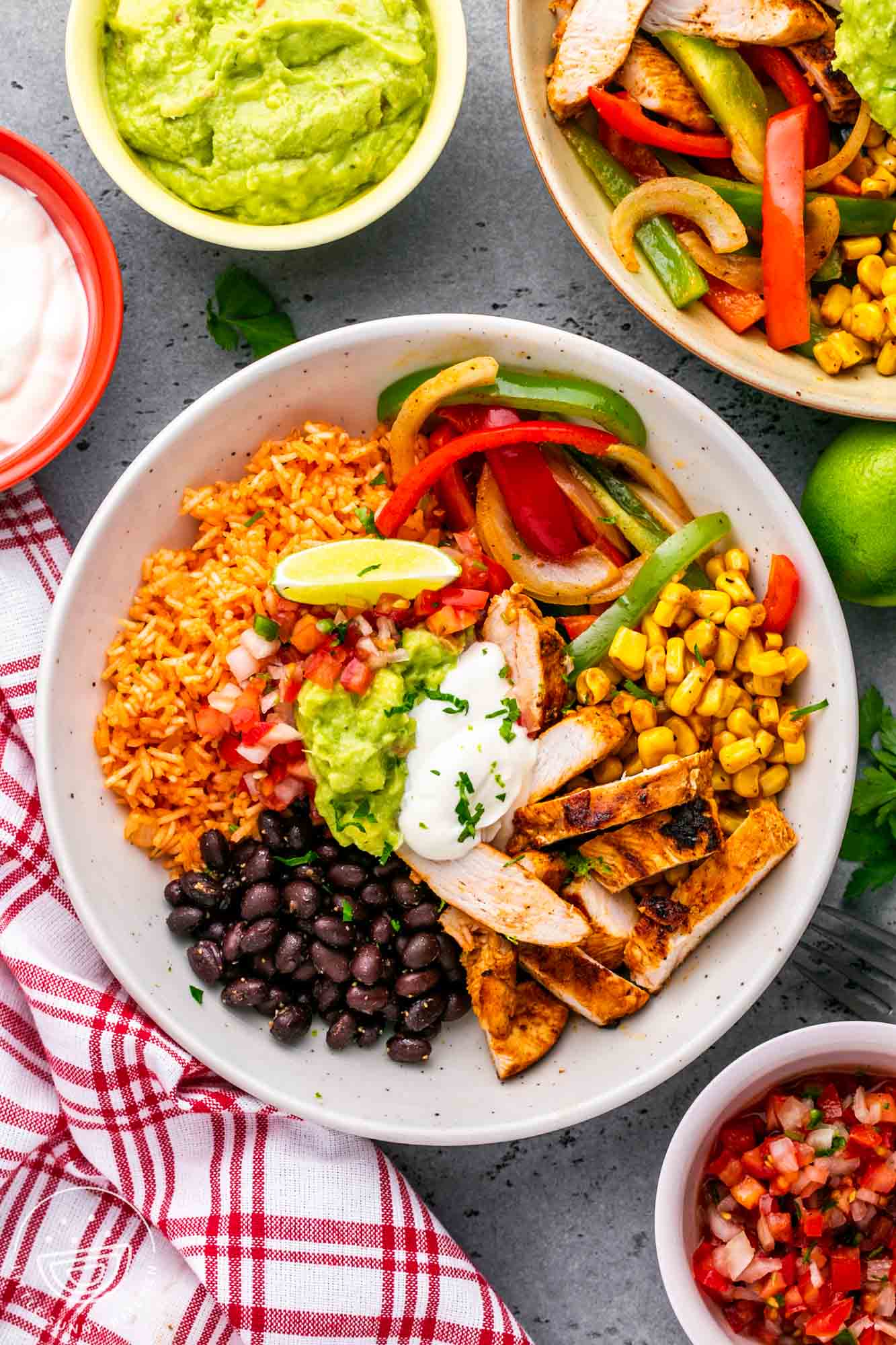 a white bowl filled with mexican rice, sliced chicken, fajita veggies, corn, and beans. Topped with sour cream and guacamole.