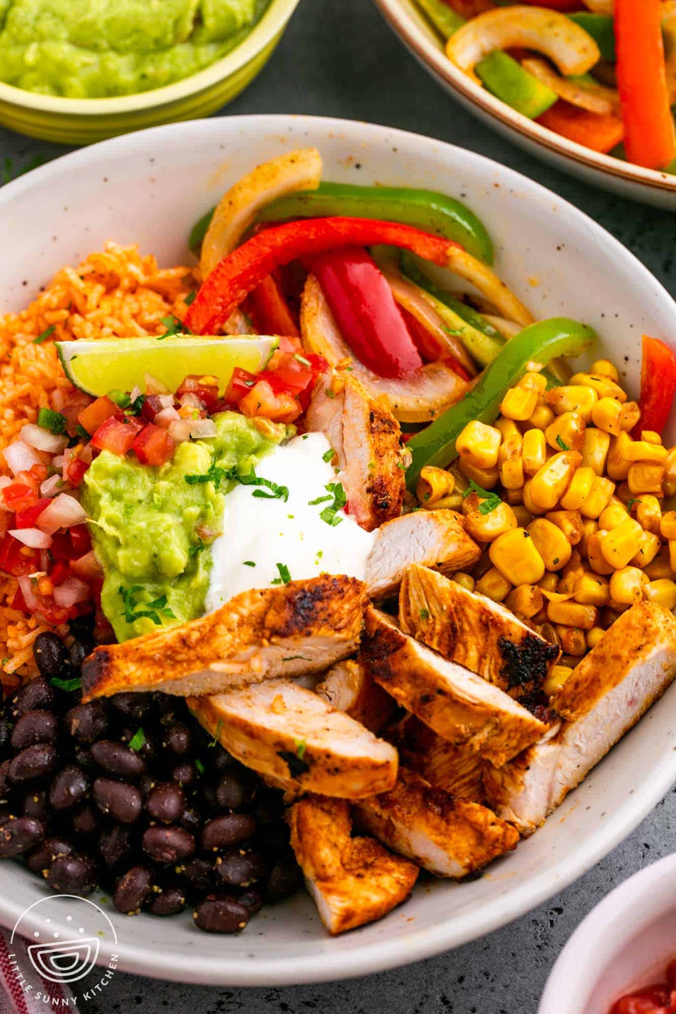 a white bowl filled with mexican rice, sliced chicken, fajita veggies, corn, and beans. Topped with sour cream and guacamole. 