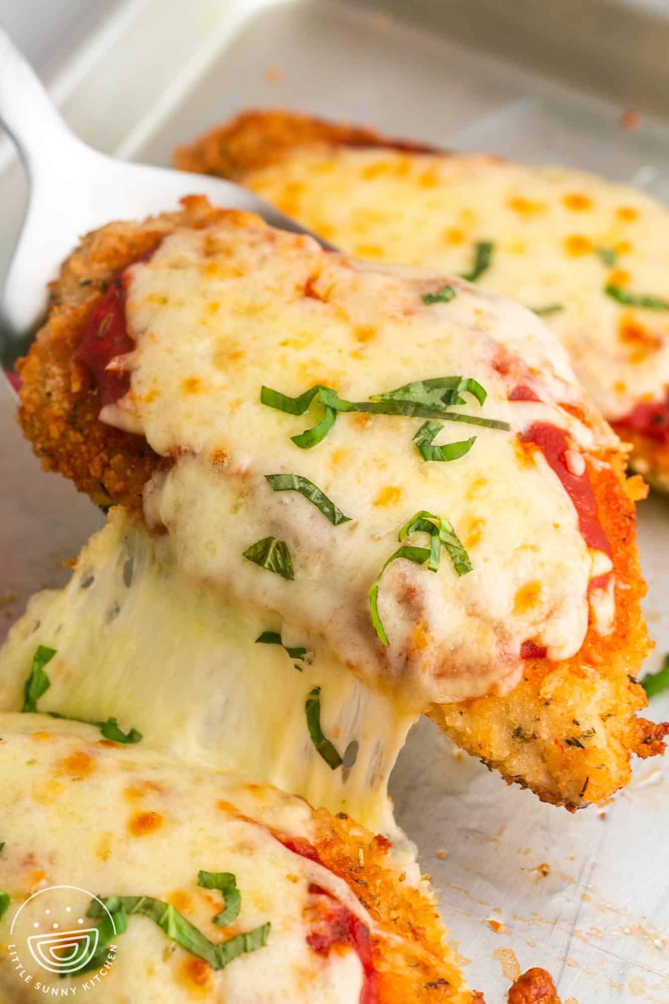 Chicken parmesan cutlets on a sheet pan, topped with sauce, cheese, and chopped basil. A spatula is lifting one off the pan, the mozzarella cheese is stringy.