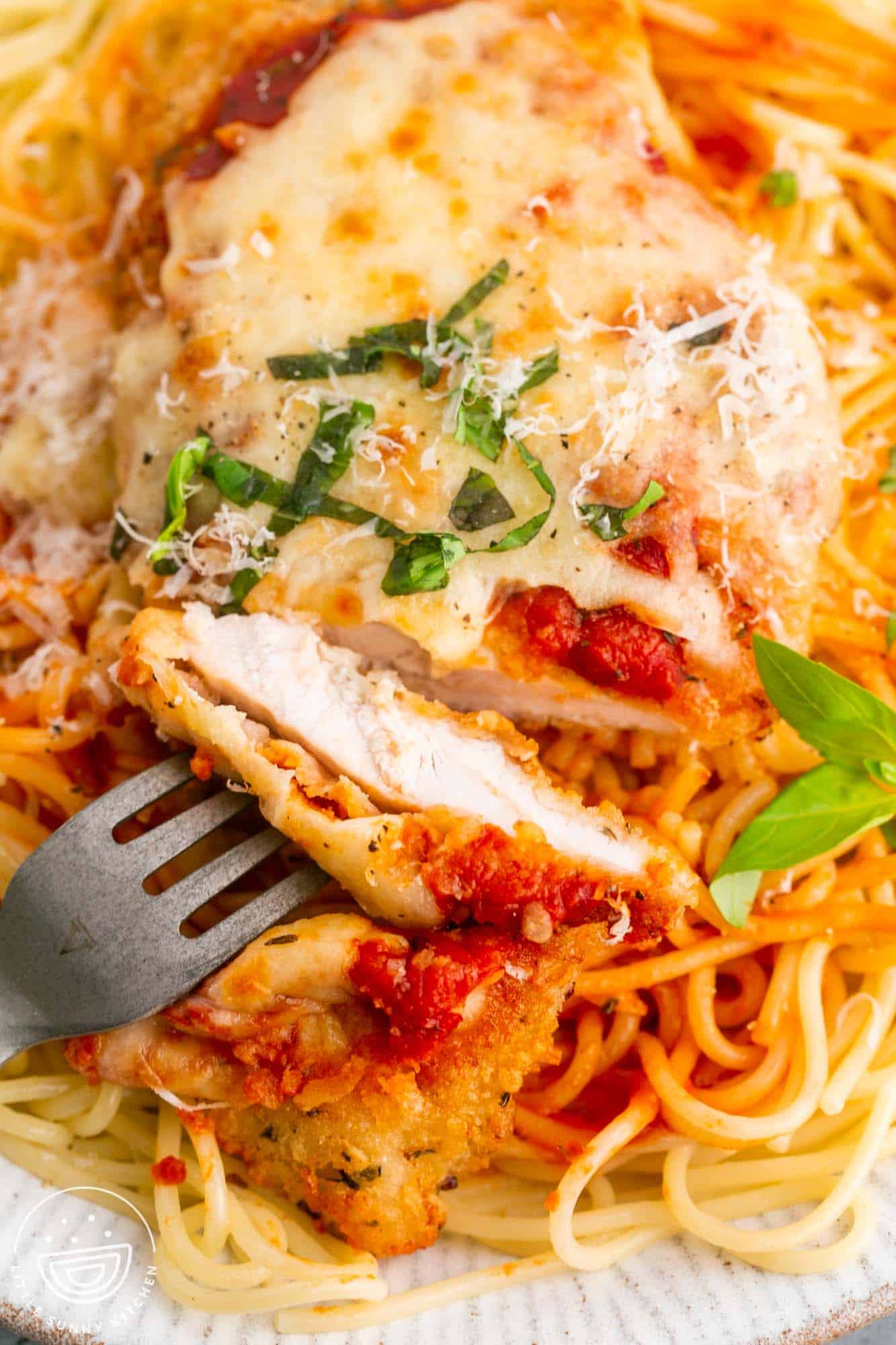 a plate of spaghetti topped with chicken parmesan. A fork is holding a slice of chicken so that you can see the internal texture. 