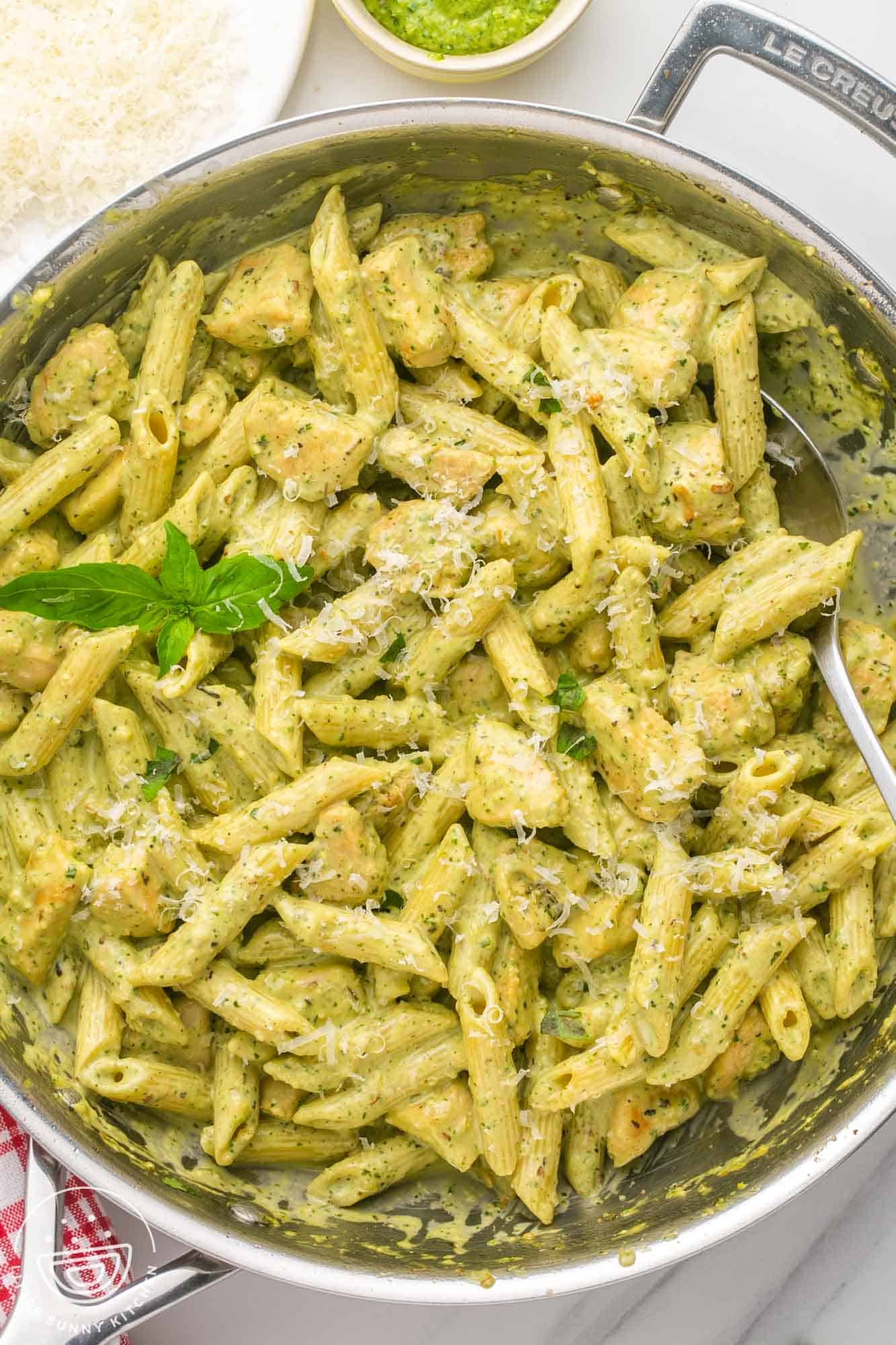 A two handled skillet filled with creamy pesto penne with chicken. A sprig of basil is added for garnish.  