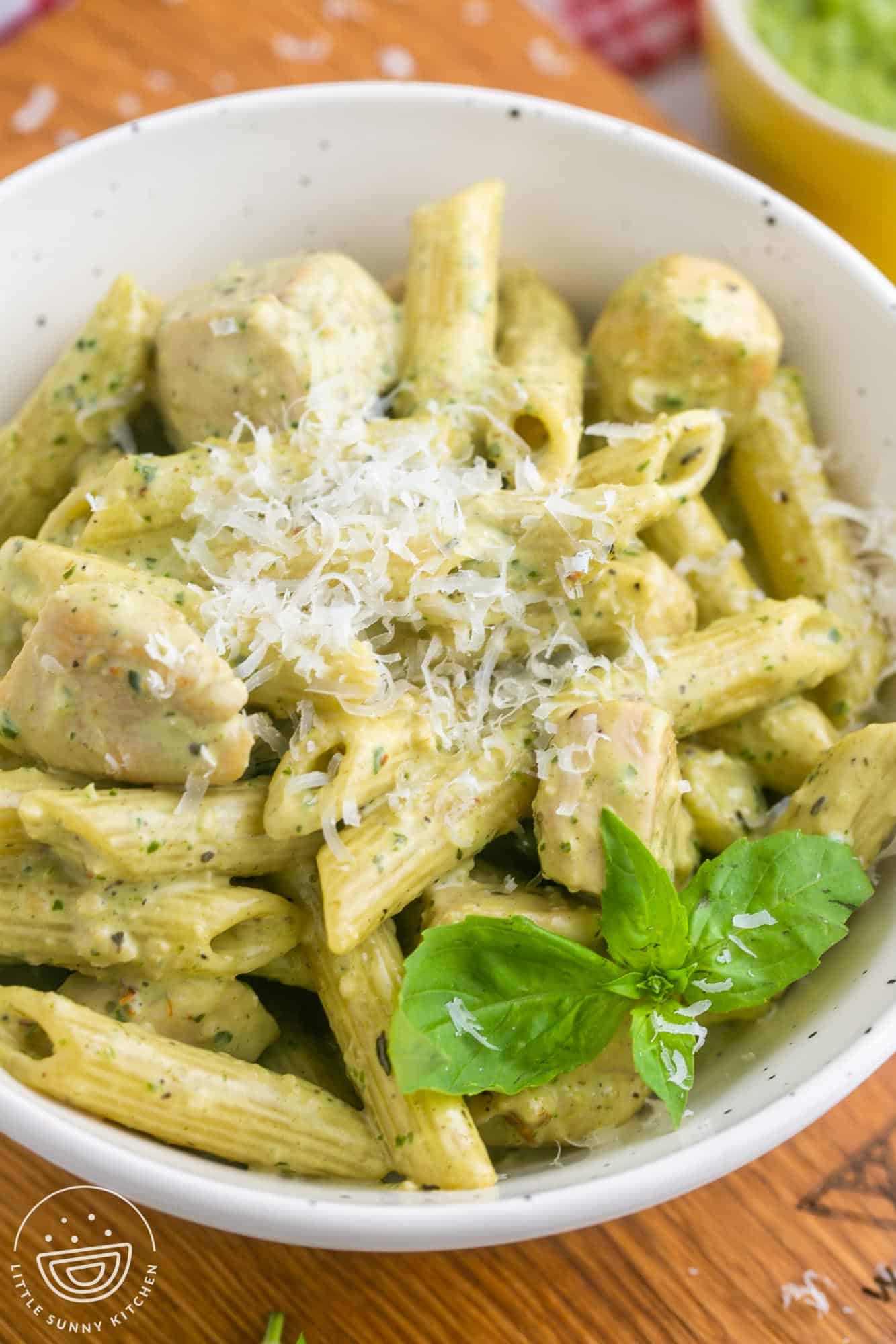 a white bowl filled with a single serving of creamy chicken pesto pasta. It's made with penne, topped with grated parmesan cheese and a sprig of fresh basil.