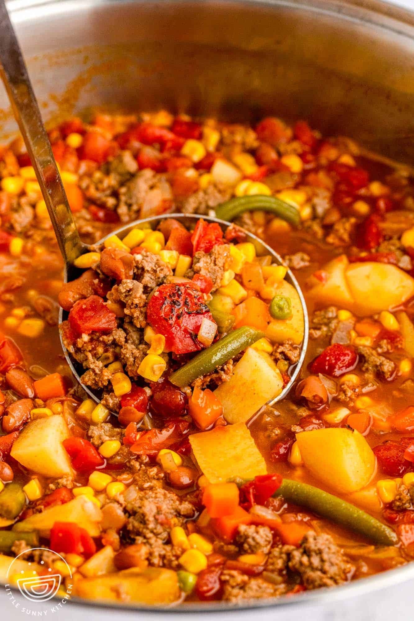closeup view of a metal pot of cowboy soup with fire roasted tomatoes and corn. A ladle is lifting up a serving to show the detail.
