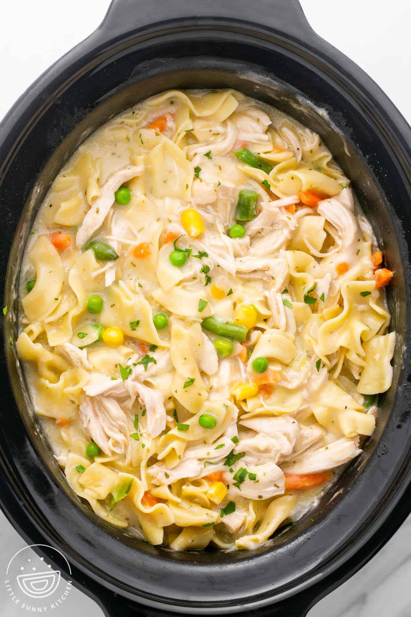 A black crockpot filled with creamy noodles with mixed veggies and shredded chicken. Viewed from overhead.