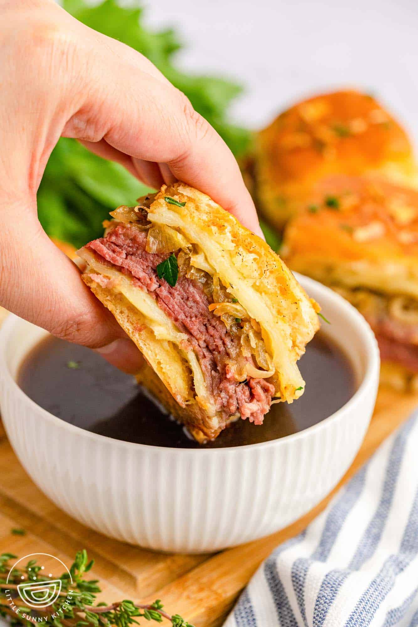 a hand dipping a french dip slider into a bowl of au jus