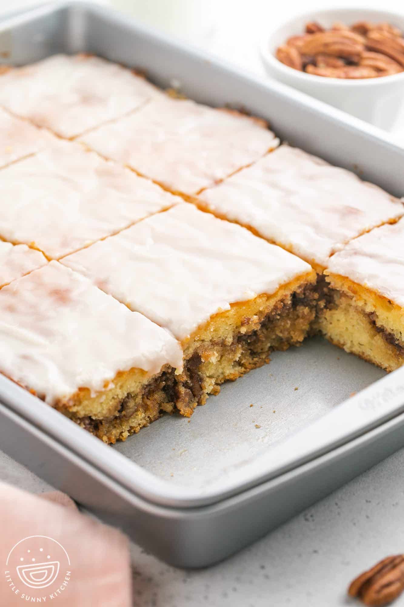 a metal cake pan of honey bun cake with icing. the cake is cut into squares, and two have been removed.