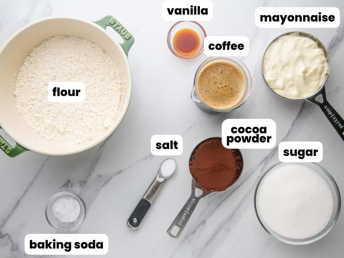 the ingredients needed to make old fashioned mayonnaise cake, all in separate bowls and measuring spoons. Each ingredient is labeled with a white text box. 
