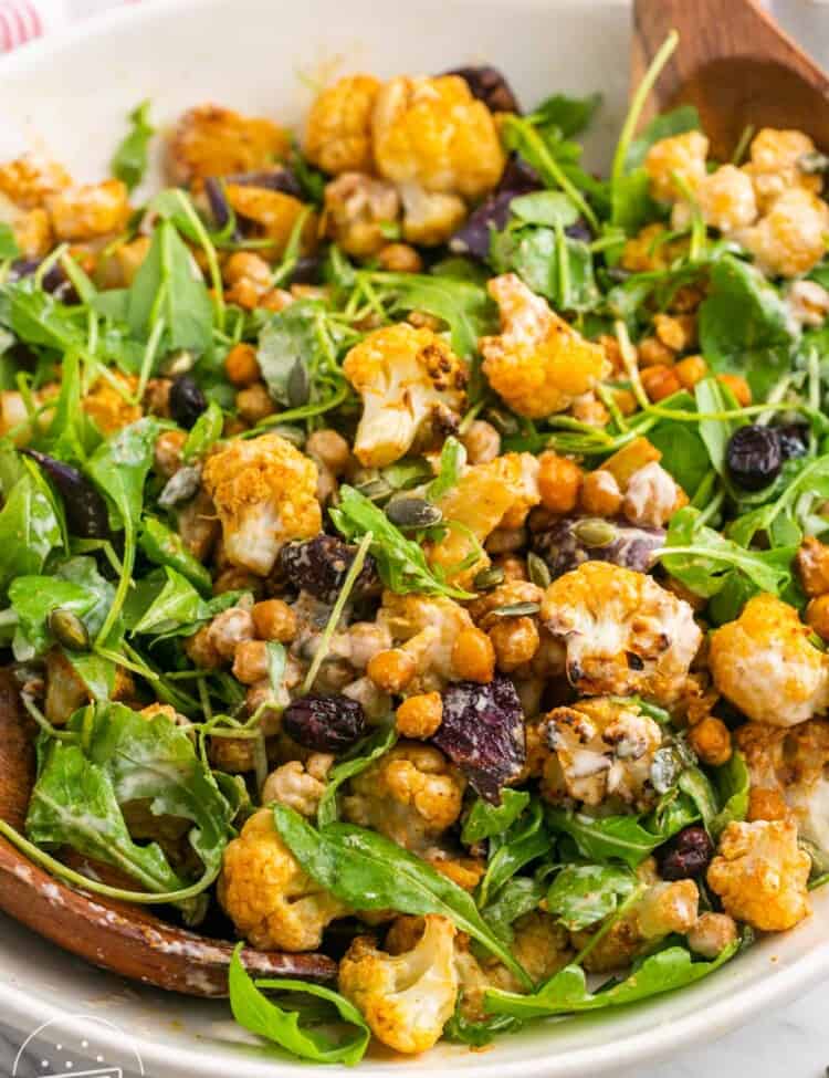 Roasted cauliflower salad with arugula in a large white bowl, and wooden servers on the sides.