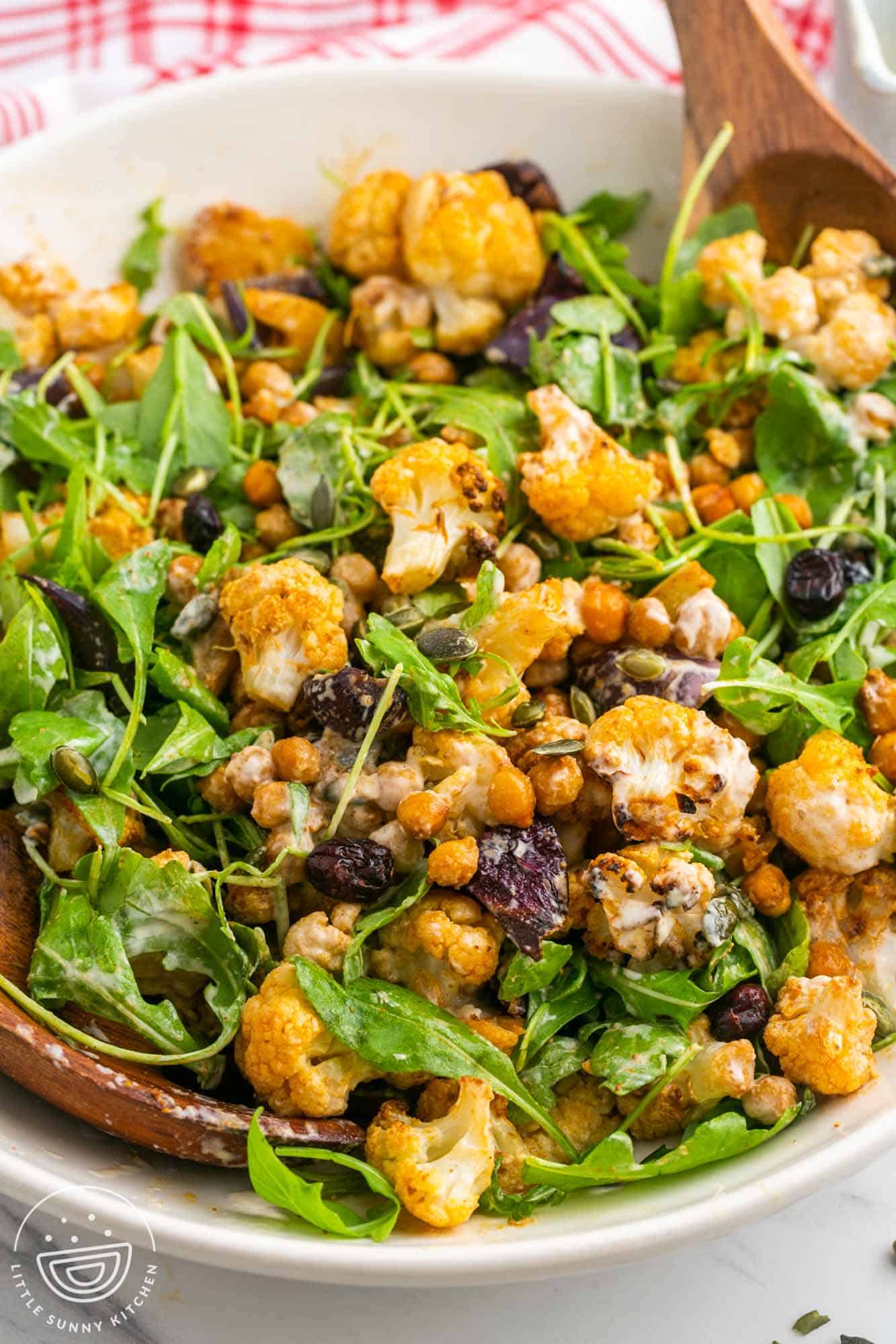 Roasted cauliflower salad with arugula in a large white bowl, and wooden servers on the sides.
