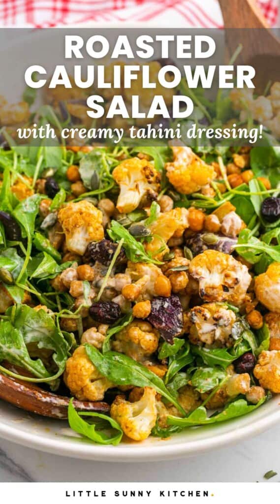 Roasted cauliflower salad with arugula in a large white bowl, and wooden servers on the sides. With overlay text that says "roasted cauliflower salad with creamy tahini dressing"