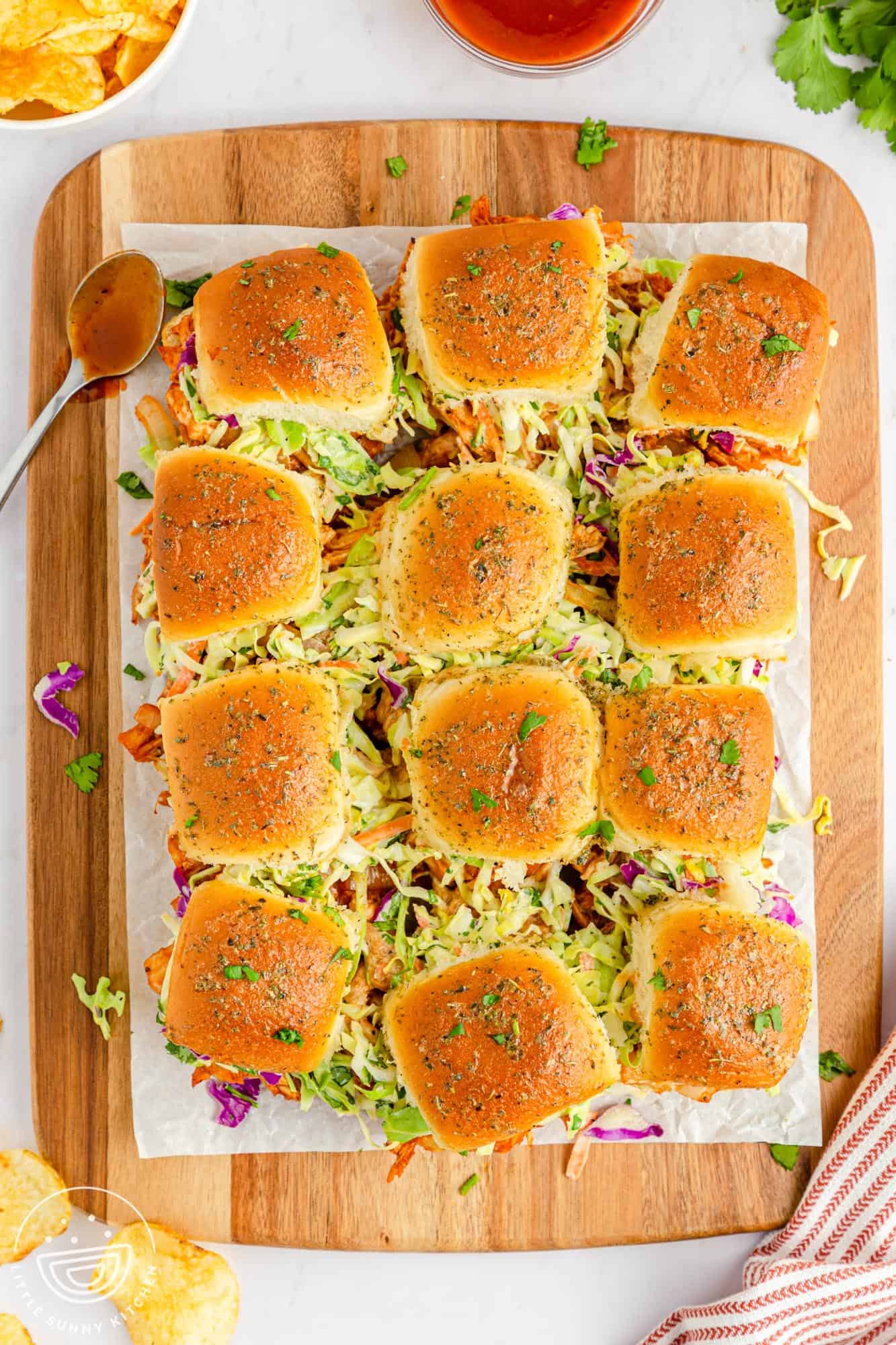 a wooden cutting board holding 12 chicken sliders on hawaiian rolls topped with herbs.