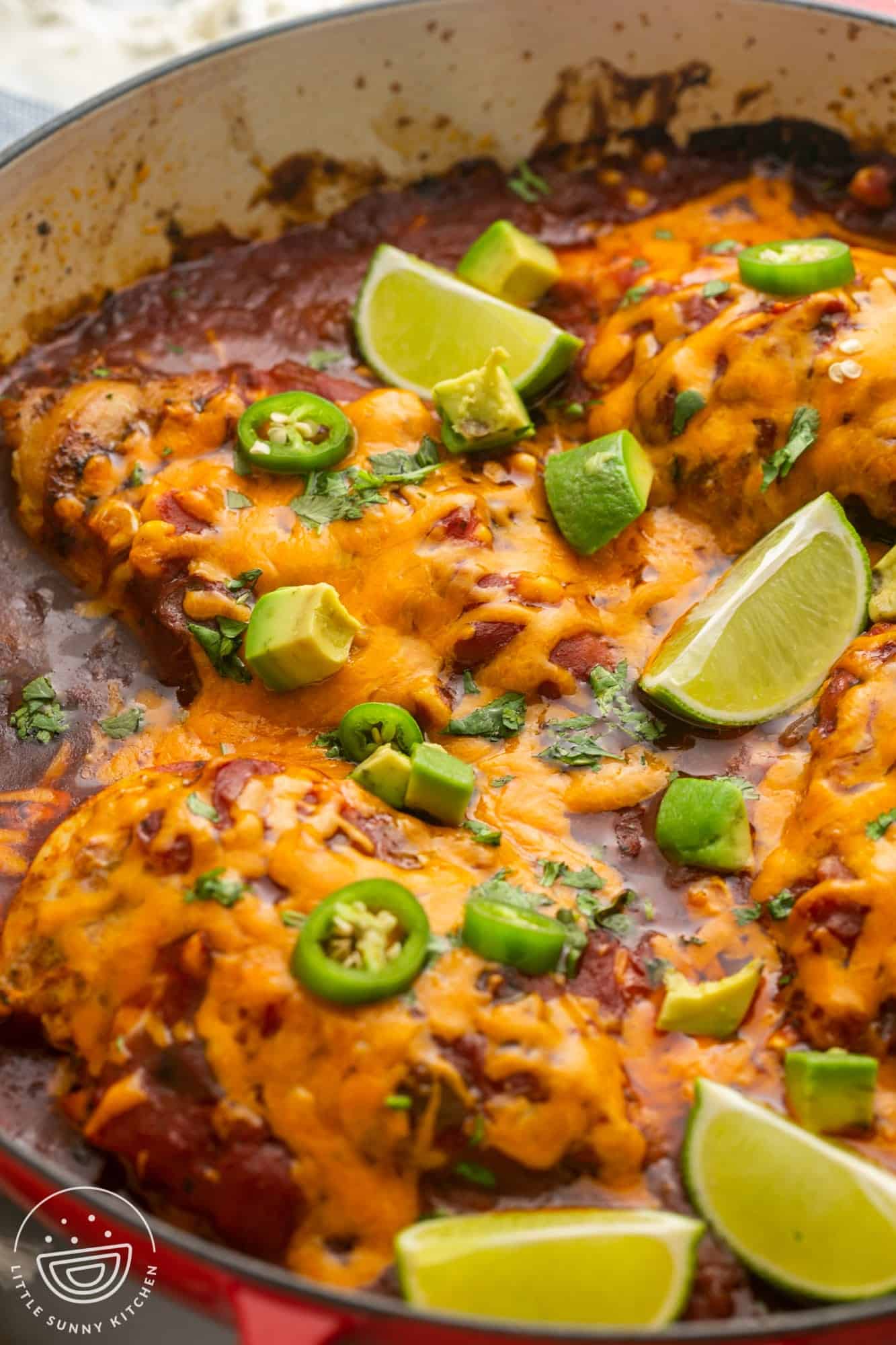 a pan of baked chicken with salsa, topped with cheese, diced avocado, lime wedges.