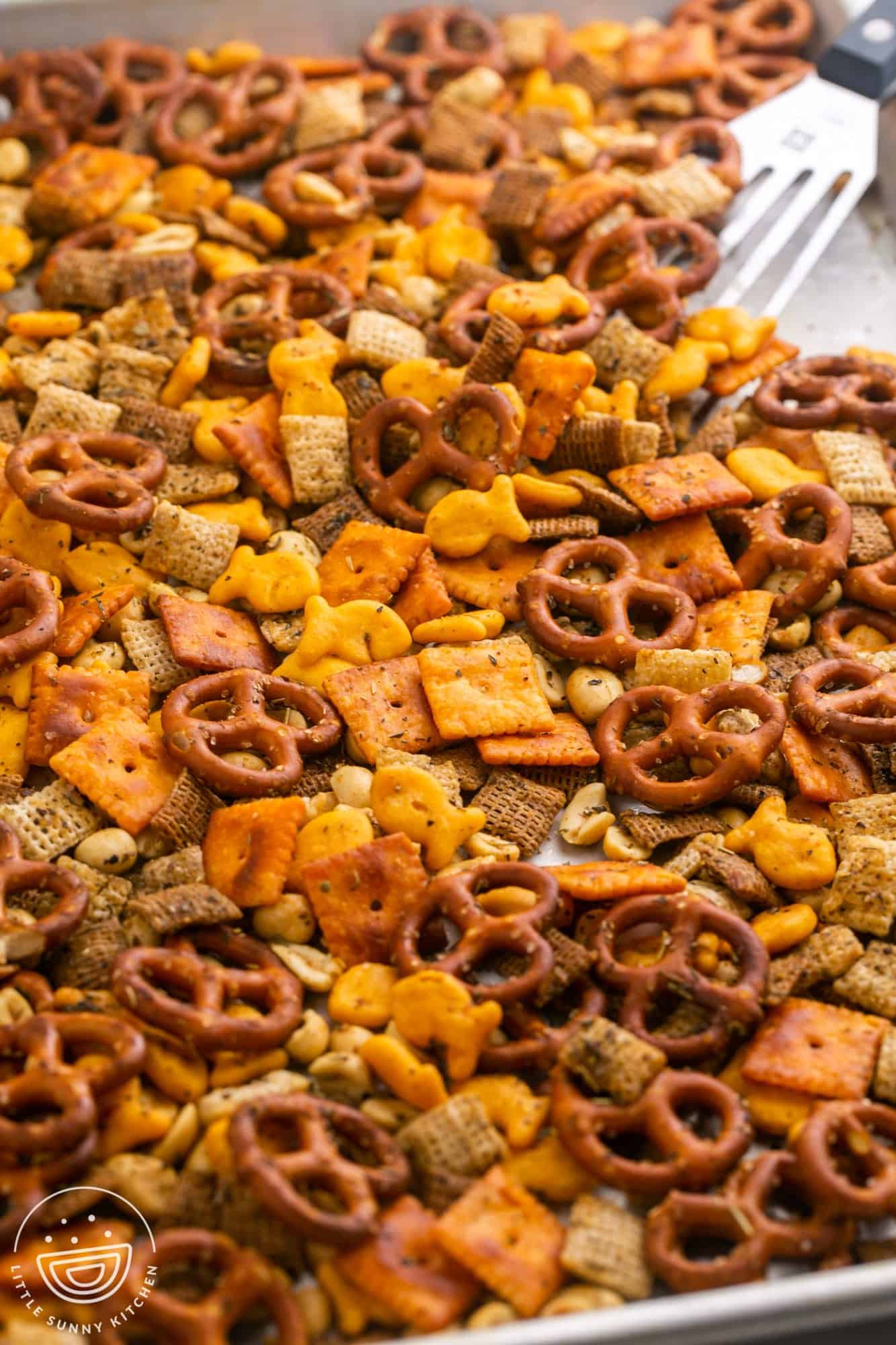 chex mix with pretzels, cheeze its, goldfish, and peanuts on a baking sheet, a spatula is stirring them.