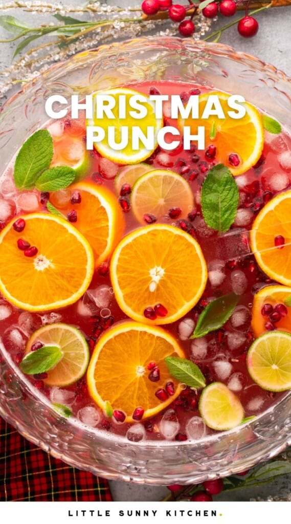 Overhead shot of a punch bowl filled with cranberry punch, sliced orange, lime slices, ice, mint leaves and pomegranate seeds. And overlay text that says "Christmas Punch"