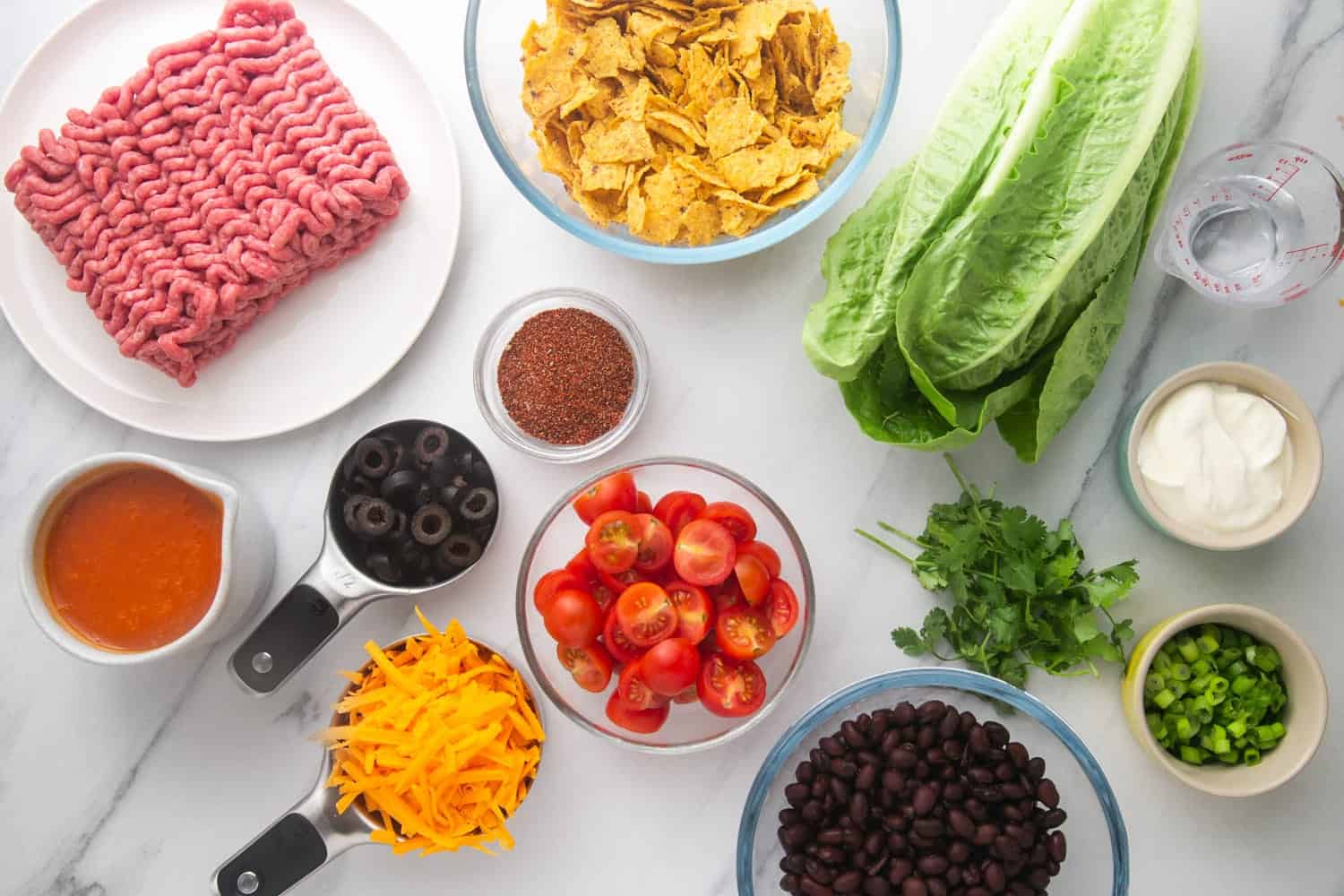 Ingredients needed for making Frito taco salad including ground beef, black beans, lettuce, cherry tomatoes, cheese, spices, and doritos.