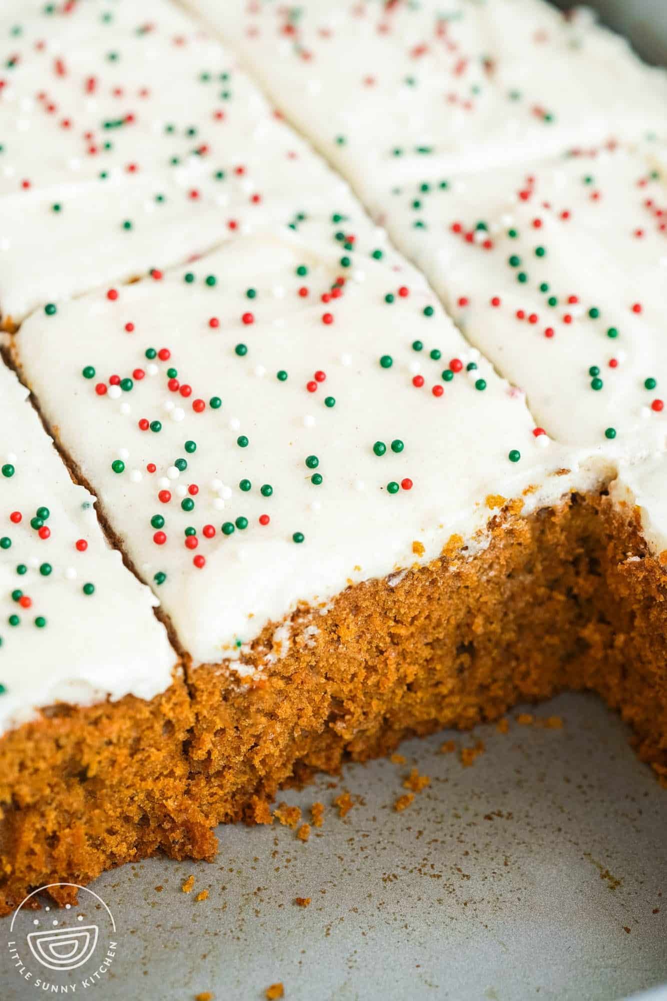a frosted gingerbread cake with sprinkles in a metal cake pan. Two slices have been removed, and the rest of the cake is cut in squares.