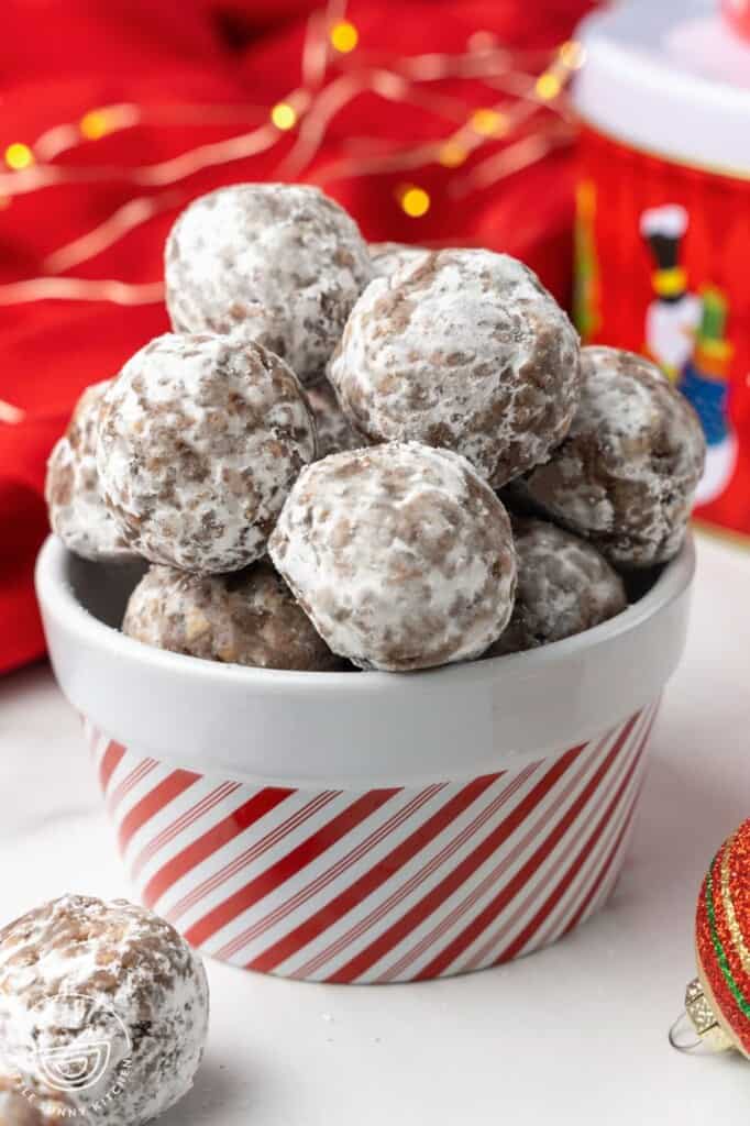 Rum balls rolled in powdered sugar, piled into a candy cane striped bowl. Christmas lights are seen in the background of the image.