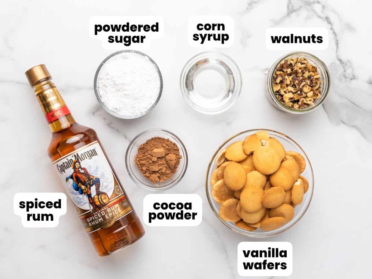 The ingredients needed to make rum balls, including captain morgan spiced rum, nilla wafers, and walnuts, in separate bowls, arranged on a counter.