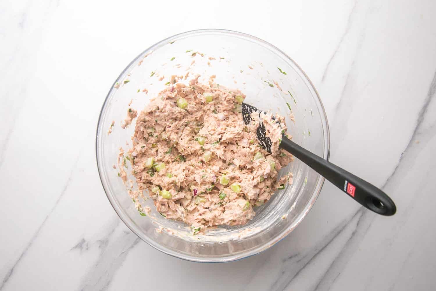 a glass bowl filled with tuna salad that is being stirred with a spatula.