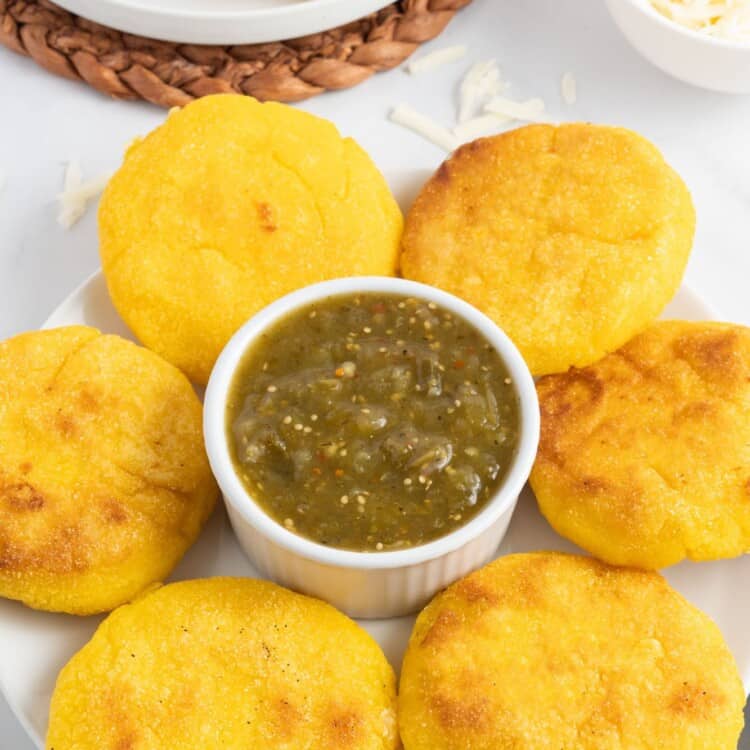 a large plate with six arepas con queso surrounding a ramekin of green salsa.