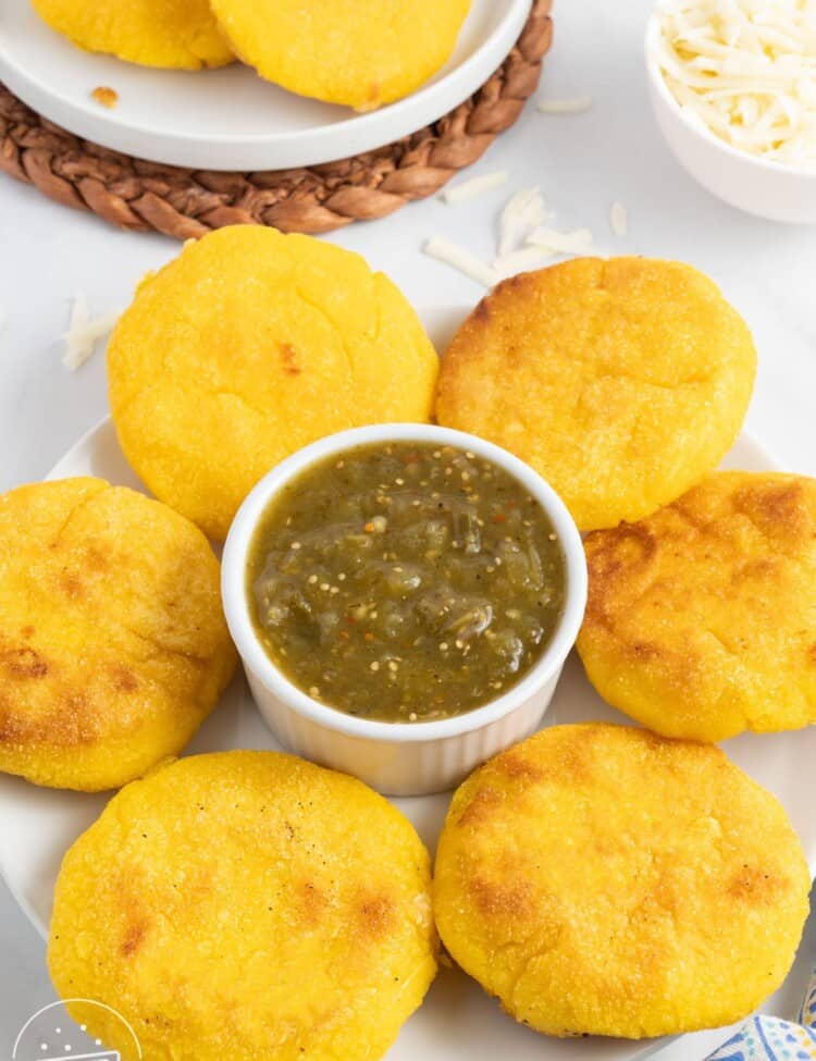 a large plate with six arepas con queso surrounding a ramekin of green salsa.