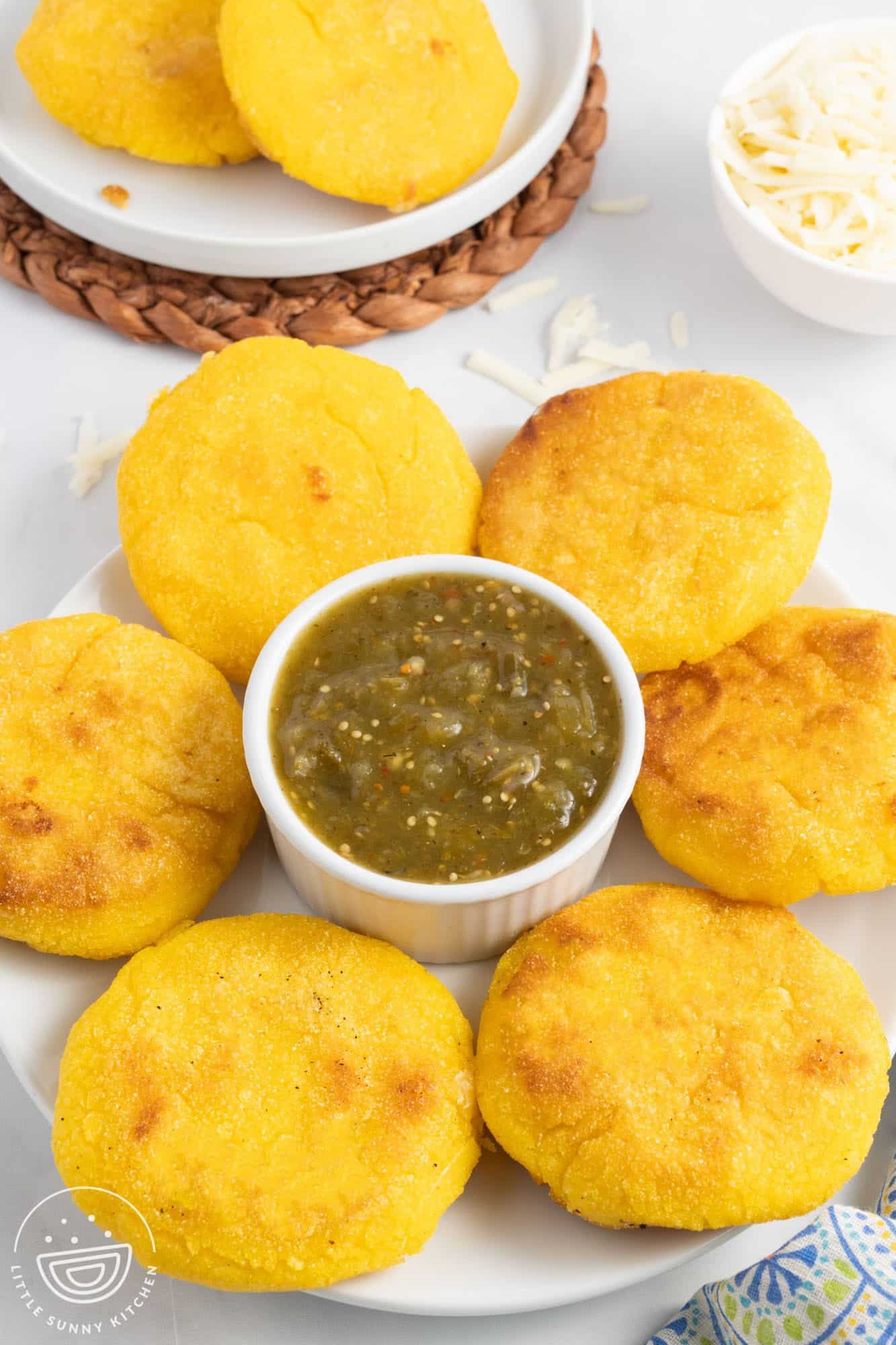 a large plate with six arepas con queso surrounding a ramekin of green salsa. 