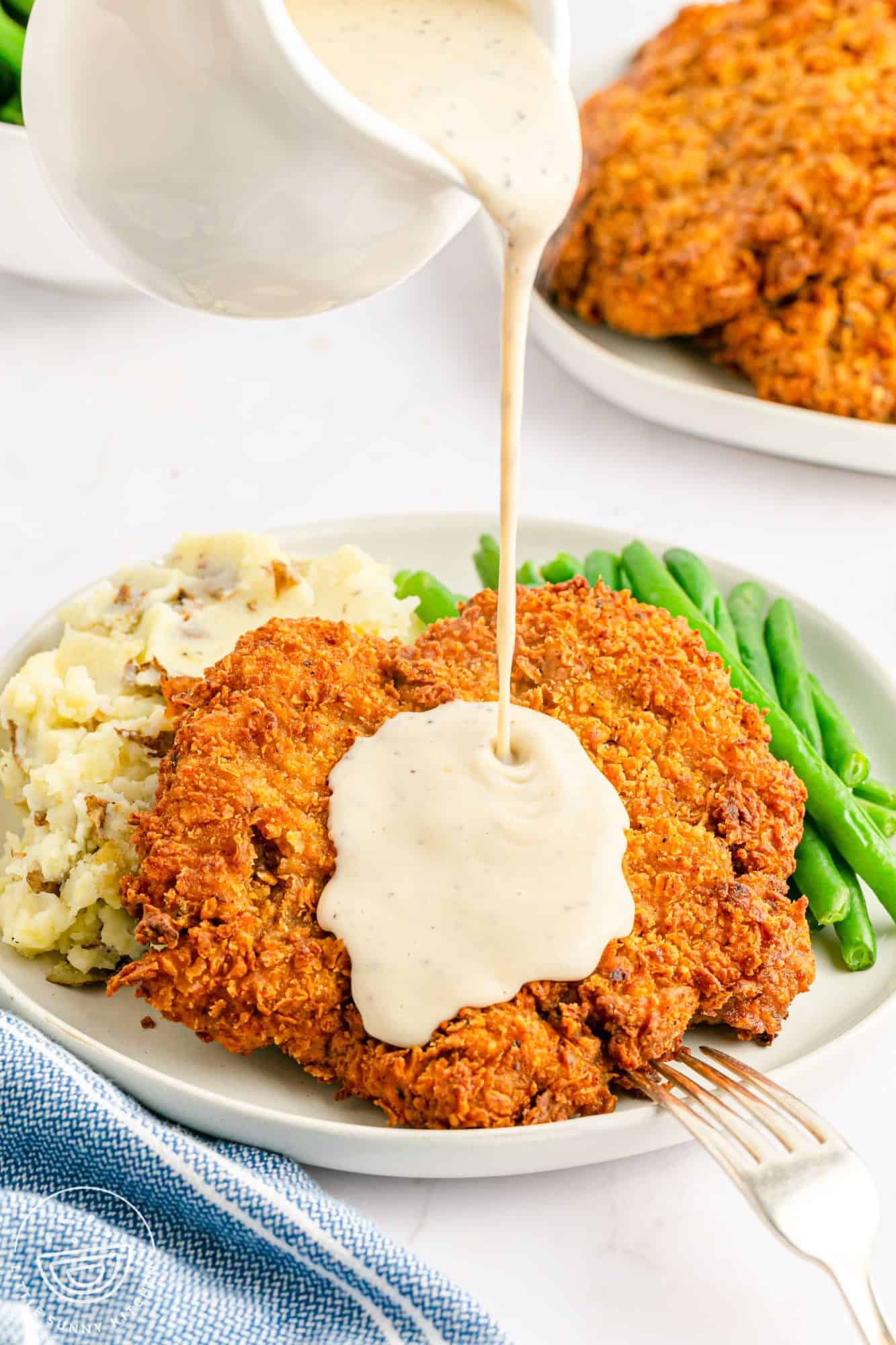 Pouring white gravy over chicken fried steak that is served on a white plate with mashed potatoes and green beans