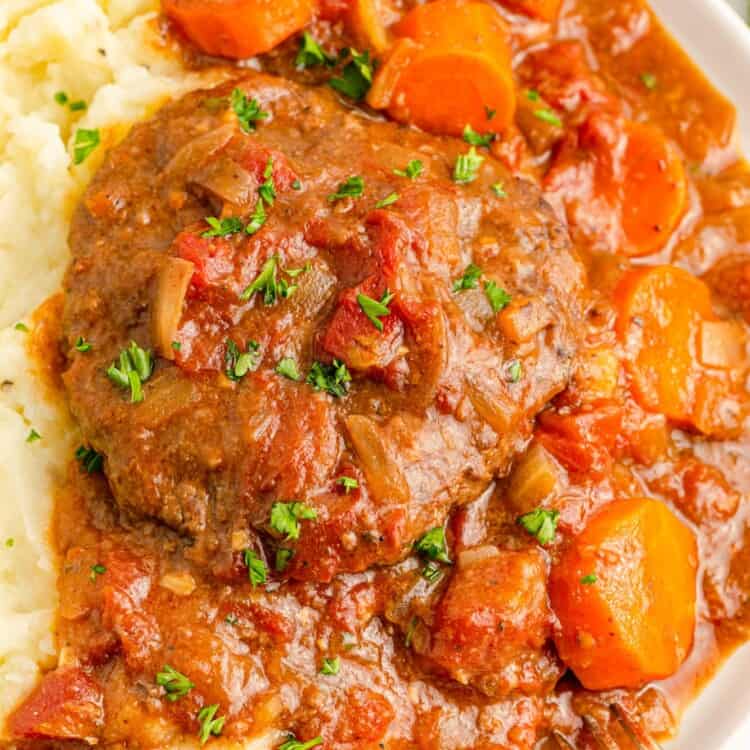Braised swiss steak with gravy, and carrots, served over mashed potatoes on a white plate.