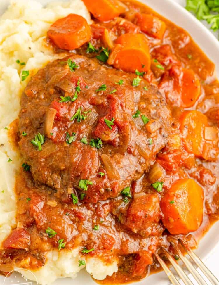 Braised swiss steak with gravy, and carrots, served over mashed potatoes on a white plate.
