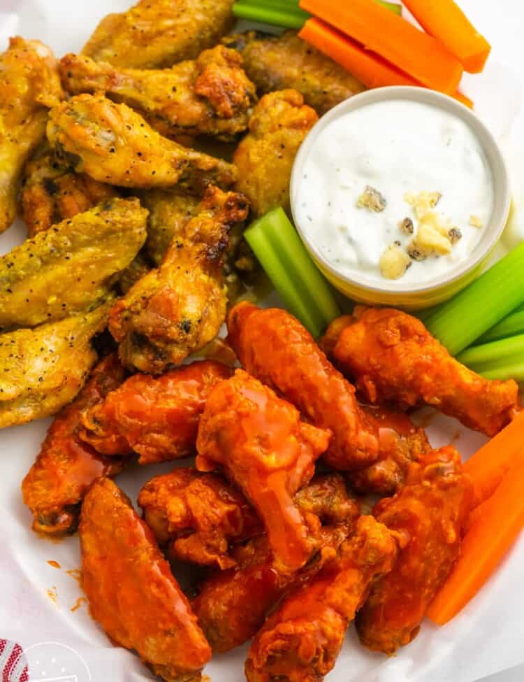 Two flavors of wings on a paper lined platter with a side of blue cheese dressing and carrot and celery sticks