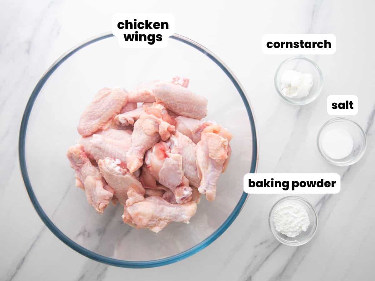 a glass bowl of raw chicken wings next to three small bowls of cornstarch, salt, and baking powder. The ingredients are each labeled with a text box overlay