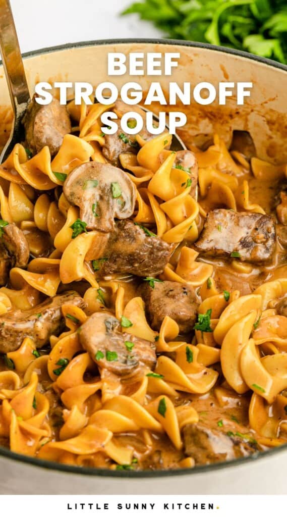 a white enameled dutch oven filled with beef stroganoff soup with lots of egg noodles, cubes of beef and sliced mushrooms.