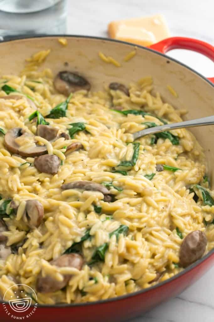 a red enameled skillet filled with creamy mushroom spinach orzo. A spoon is in the pan.